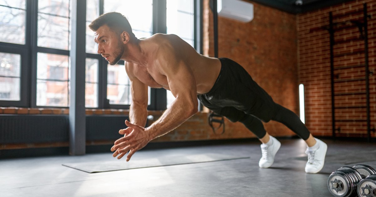 A young, fit man in black tights and without a shirt performs a clap push-up inside an empty fitness center.