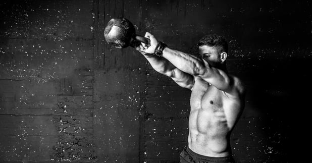A black-and-white photo of a fit young man without a shirt swingining a kettlebell with both hands for a workout.